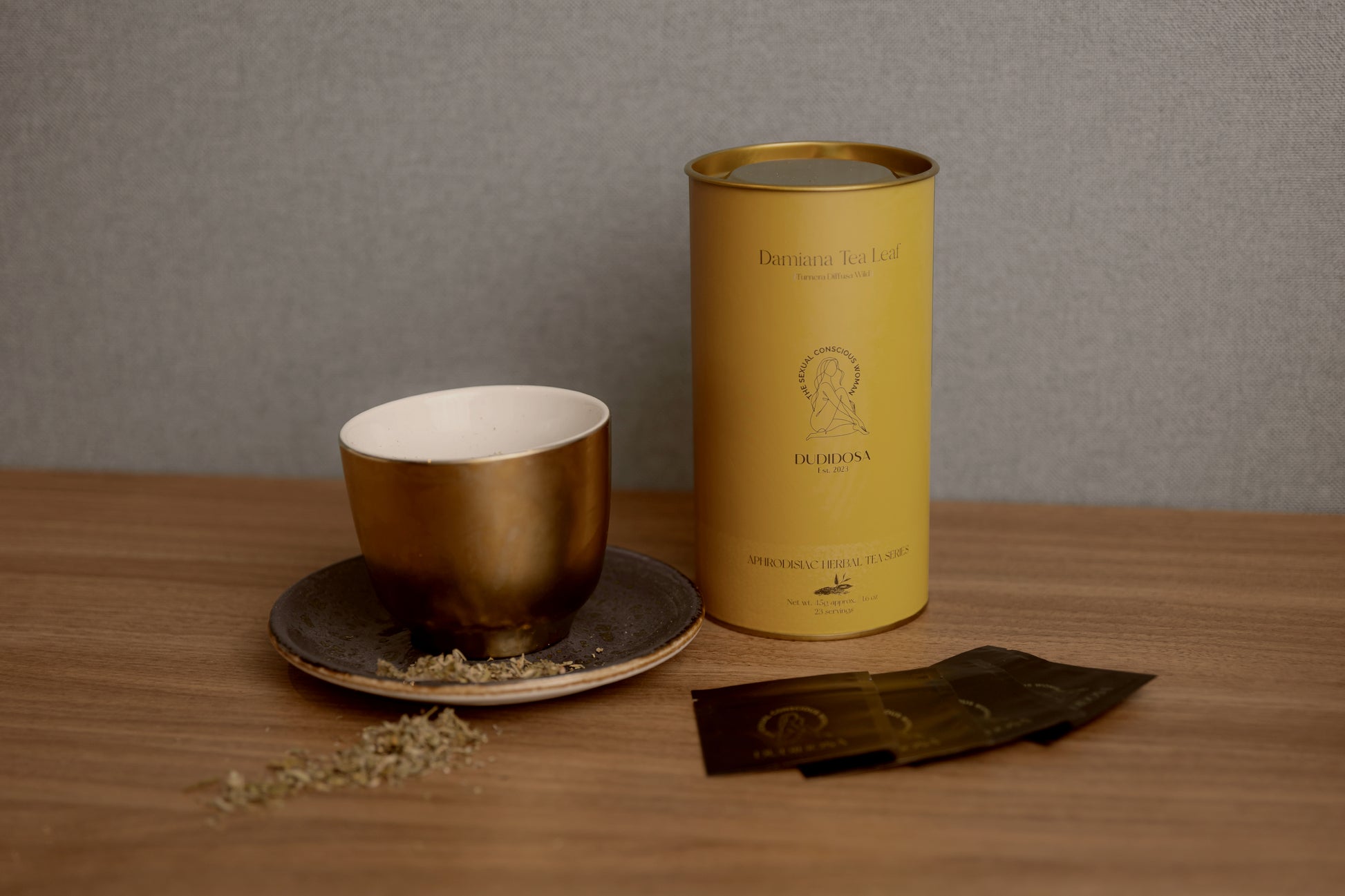 DUDIDOSA Damiana Tea Leaf tin placed beside a golden teacup on a ceramic saucer, with loose tea leaves scattered on a wooden table. The warm, minimalist setting reflects DUDIDOSA’s calming, sensual ritual — a natural herbal tea crafted to ease the mind and awaken connection.