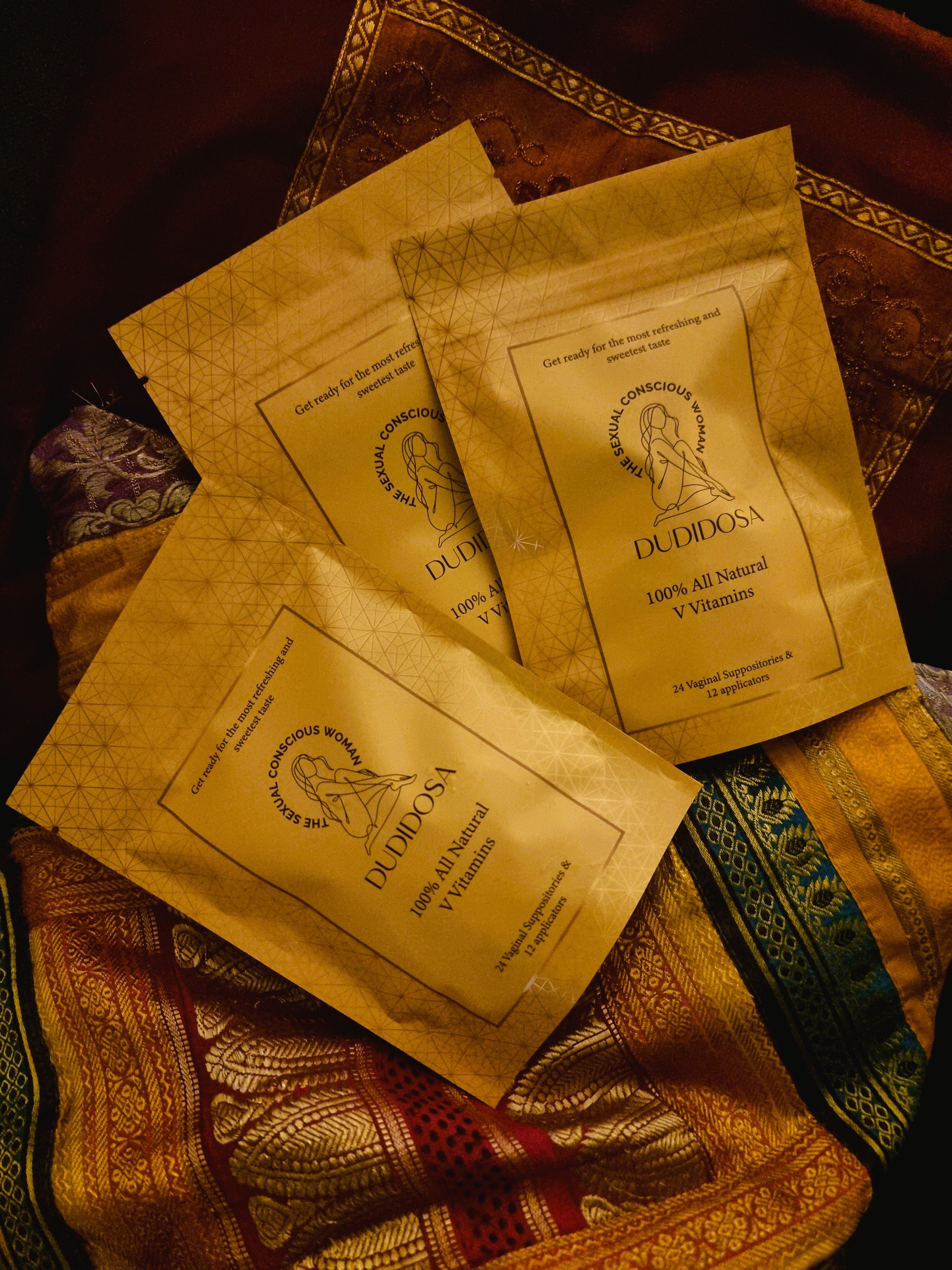 Three golden DUDIDOSA V-Vitamins pouches resting on richly patterned silk fabric in warm tones of red, gold, and green. The scene evokes a sensual, luxurious atmosphere —reflecting the product’s natural promise to balance pH, support freshness, and subtly sweeten taste for intimate wellness.